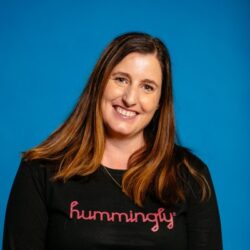 Smiling woman with long brown hair wearing a black hummingly shirt, posing against a blue background—channeling the resilience of Jolie Wills after securing a rebuild permit.