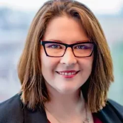Kate Bulger, a woman with brown hair and glasses, smiles in a bright setting while wearing a black blazer and red top—ready to discuss your rebuild permit needs.