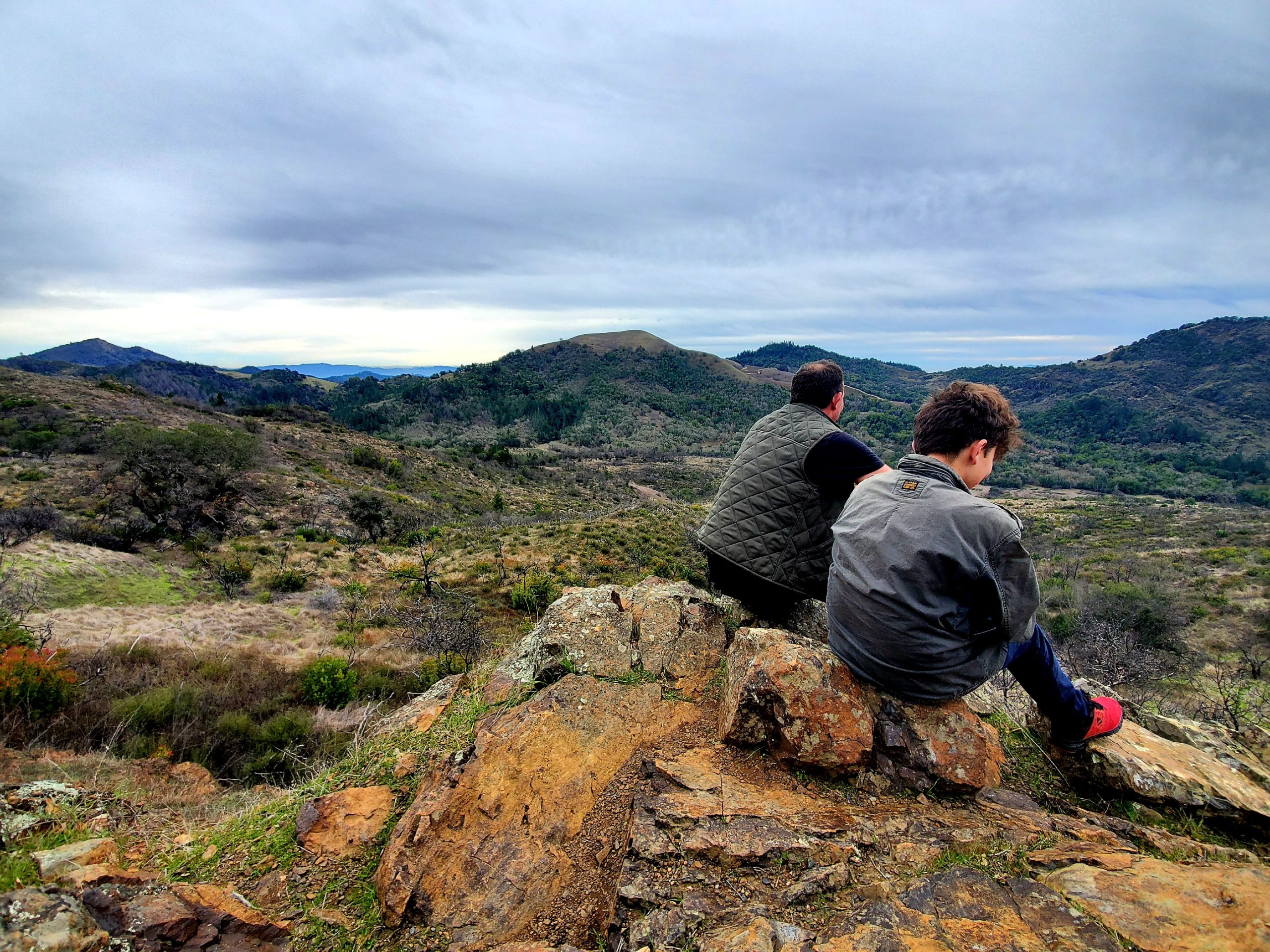 Man and his son with backs to camera sitting on rocks looking out on a mountain vista