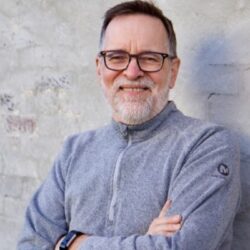 Smiling man with glasses and gray beard, possibly Michael Morter, wearing a gray sweater stands with arms crossed in front of a light wall—ready to discuss your rebuild permit needs.
