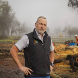 Man in a vest stands on a foggy worksite with two workers in safety gear and cut logs behind him, possibly discussing a rebuild permit as overseen by Tennis Wick.