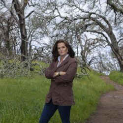 Woman with brown hair stands with arms crossed on a grassy trail surrounded by leafless trees.