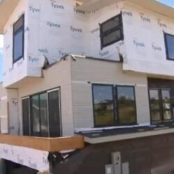 A two-story house under construction with exposed insulation wrap and unfinished exterior walls.