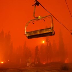 Empty ski lift and burning forest under an orange sky filled with smoke from a wildfire.
