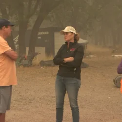 Three people stand outside talking; one wears a mask and safety vest, with smoky air and trees in the background.
