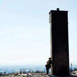 A firefighter stands by a tall brick chimney amid debris, overlooking a hazy landscape.