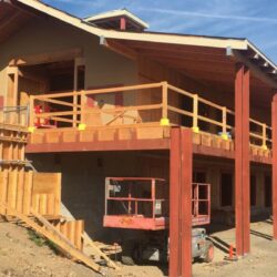 A large house under construction with wooden framing, scaffolding, and tools around the site on a sunny day.