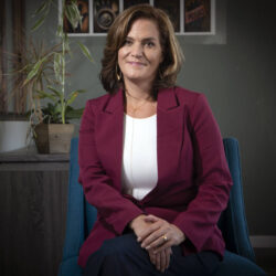 A woman in a maroon blazer sits on a blue chair, smiling, with plants and photos in the background.