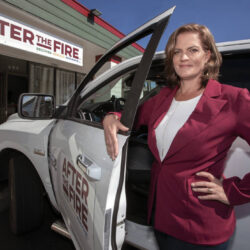 Woman in a red blazer stands by a white car with After the Fire logo, outside a matching storefront.