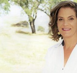 A woman in a white shirt smiles outdoors with trees and sunlight in the background.