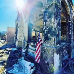A damaged, burned building with an American flag amidst rubble and bright sunlight in the background.