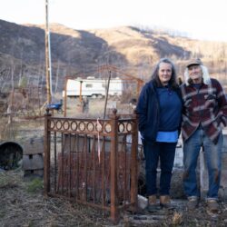 Two people stand together outdoors by a metal fence, with a dog nearby and hills in the background.