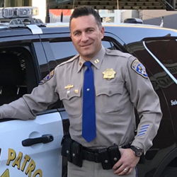 Police officer Aristotle Wolfe in uniform stands by a patrol car, smiling at the camera, proudly holding a newly issued rebuild permit.