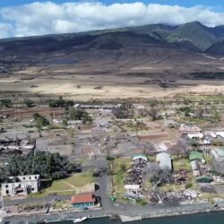 Aerial view of a coastal area with widespread fire damage, burnt buildings, and barren landscape.
