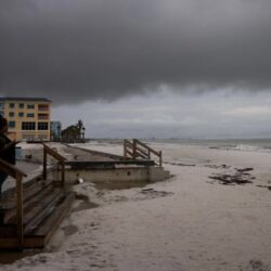 Two people stand on stairs watching a cloudy, deserted beach as another pair walks along the shore.