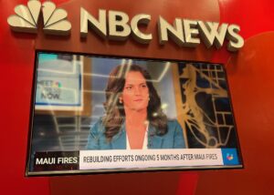 A woman speaks about Maui fire recovery on NBC News, displayed on a television with a red background.