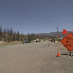 Orange Road Work Ahead sign and traffic cones on a rural road with construction vehicles in the background.