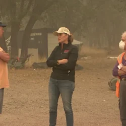 Three people talk outdoors on a smoky day; one wears a mask, another has a safety vest, and trees are in the background.