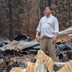A man in a white shirt stands among the charred remains of a burnt-down structure in a forested area.