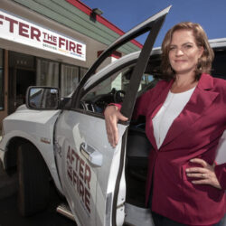 Woman in a maroon blazer stands by a white truck, in front of an AFTER THE FIRE building sign.