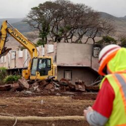 A yellow excavator demolishes a building as workers in safety gear supervise the site.