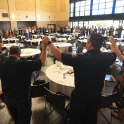 People stand in a large room, holding hands in a circle around tables, with a dog in the foreground.