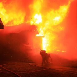 A firefighter sprays water on a house engulfed in bright orange flames at night.