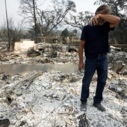 A man stands in the ruins of a burned building, wiping his face amid ash and debris.