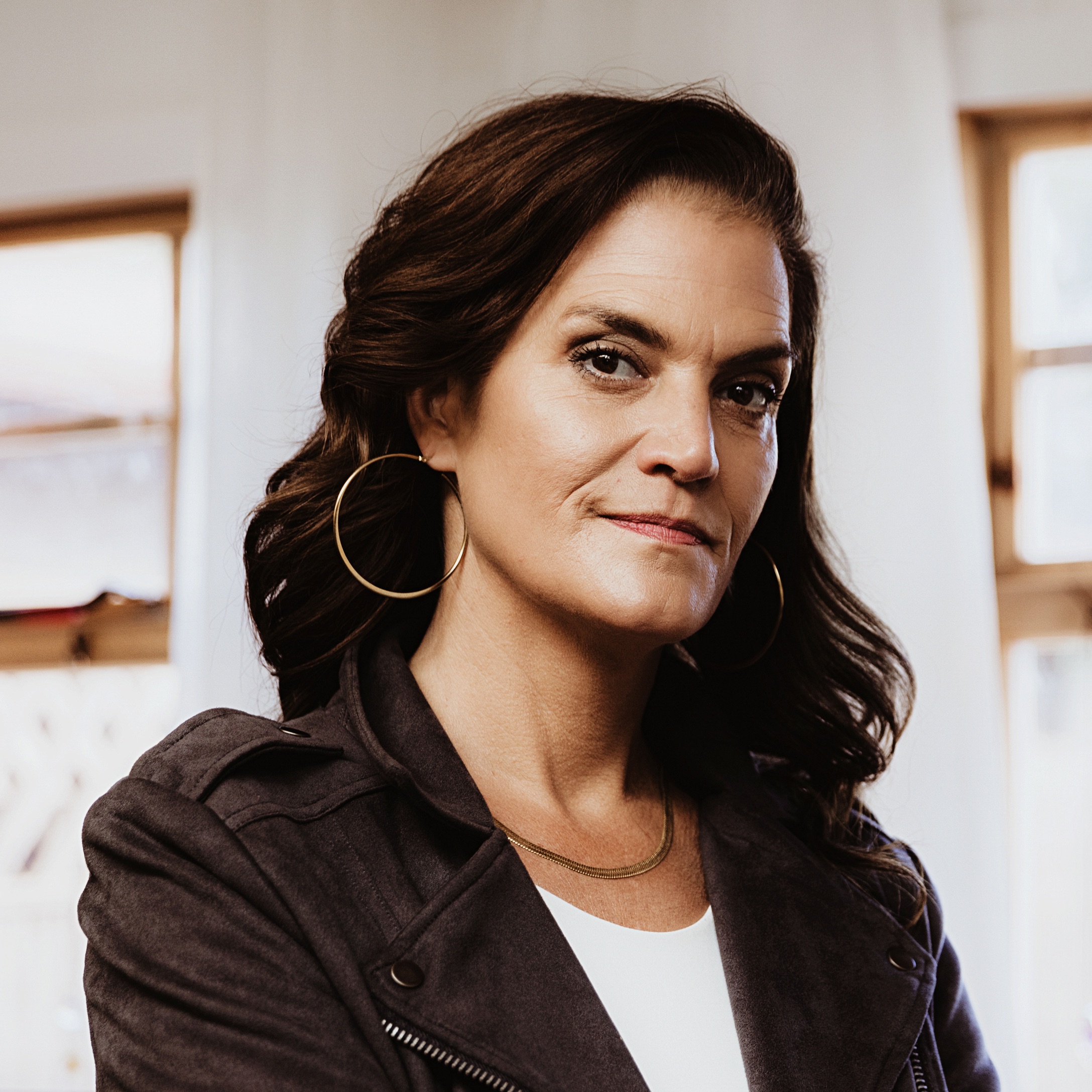 Woman with wavy dark hair, wearing hoop earrings and a dark jacket, standing indoors by a window.