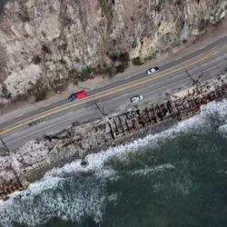 Aerial view of a coastal road with cars, rocky cliffs, and waves crashing against the shore.
