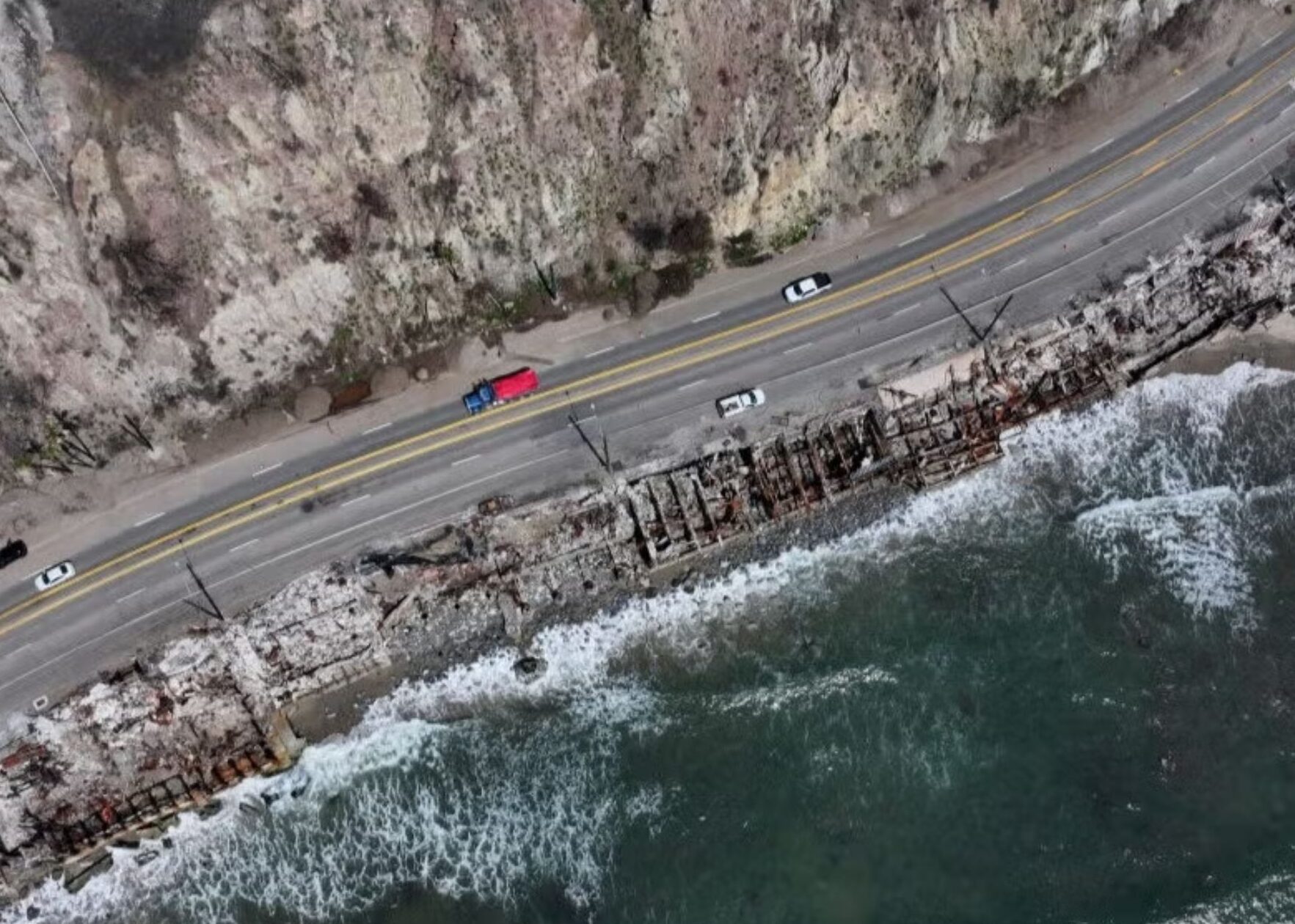 Aerial view of a coastal road with cars, rocky cliffs, and waves crashing against the shore.