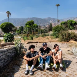 Three people sit on a stone path outdoors with mountains and trees in the background under a clear sky.