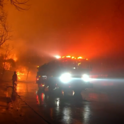 Fire truck with headlights on and firefighter respond to large blaze at night, smoke and flames fill the dark sky.