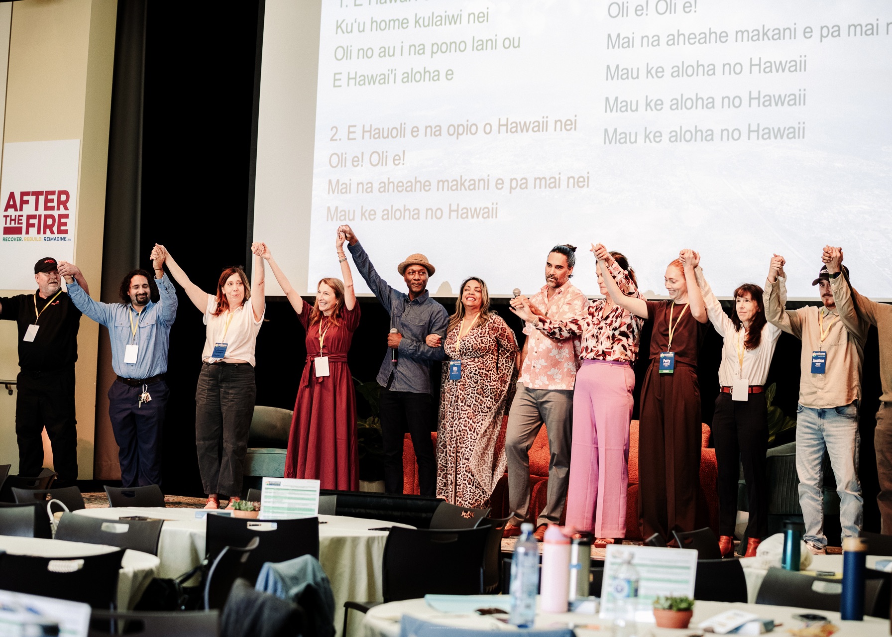 A diverse group stands on stage holding hands, smiling, in front of a screen displaying Hawaiian song lyrics—embodying community recovery and unity after the fire.