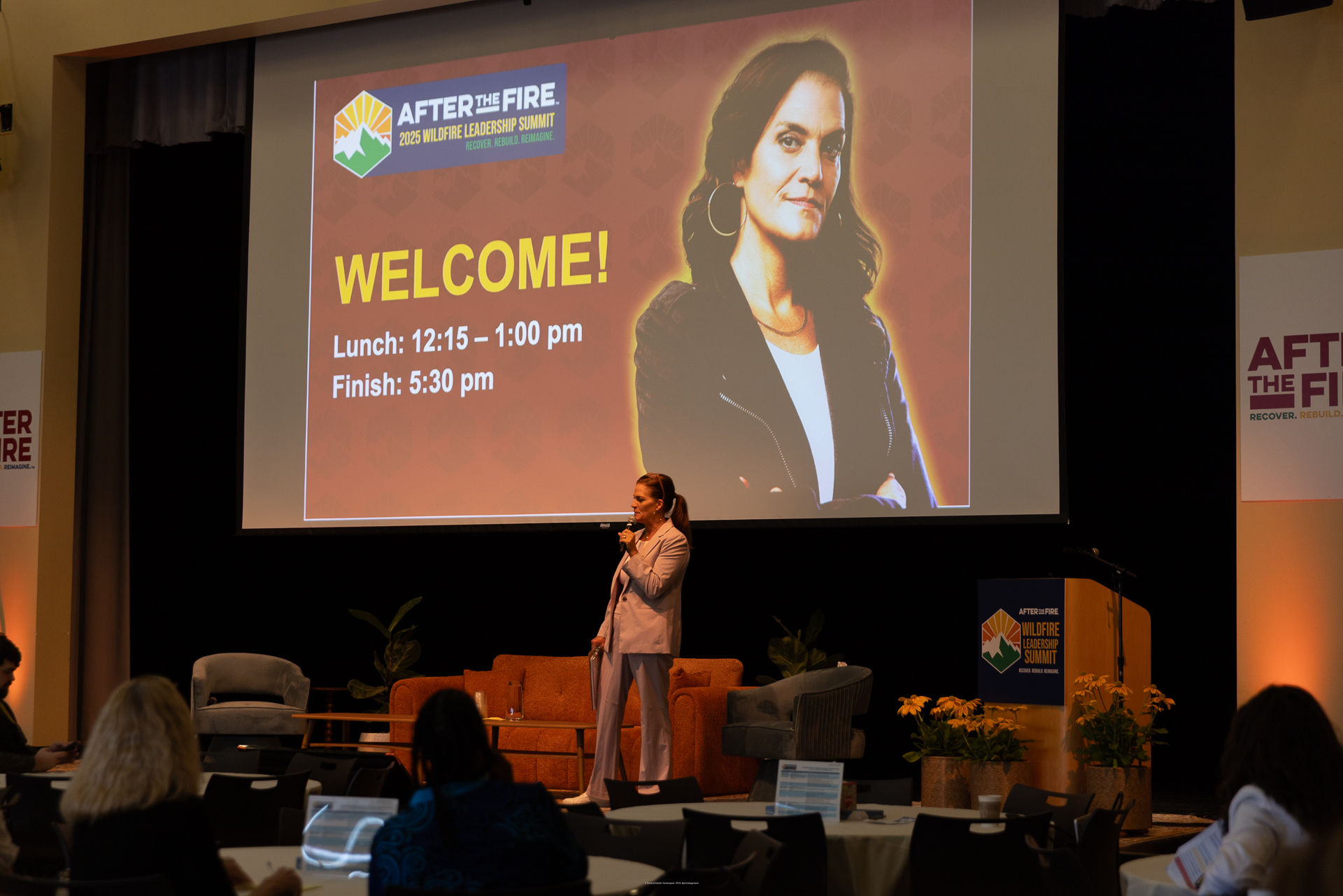 A woman speaks on stage at a leadership summit, with a welcome slide and her photo displayed behind her.