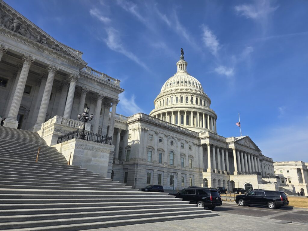 The U.S. Capitol building with its dome and steps under a blue sky, several black vehicles parked in front—partner with us to support democracy at its heart.