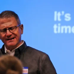 A man speaks in front of a blue screen displaying the words “It’s time.”.