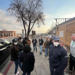 A group of people stand on a sidewalk near a construction site, some wearing masks and hats, talking together.