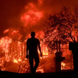 A person stands in silhouette watching a large building engulfed in flames at night.