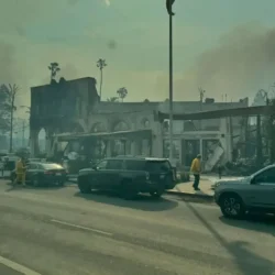Smoky scene of burned buildings, firefighters, and parked cars on a street after a fire.