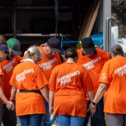 A group wearing orange Samaritans Purse shirts stands in a circle, holding hands and praying.