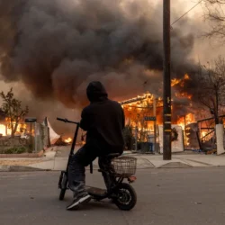 A person on a scooter watches large flames and thick smoke engulf houses on a residential street.