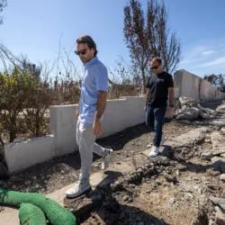 Two men walk through a burned, damaged area with debris, charred trees, and sparse vegetation.
