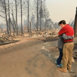 Two people embrace on a street, surrounded by the ruins of homes and charred trees after a wildfire.