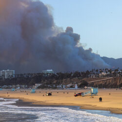 Smoke billows from a wildfire behind buildings near a sandy beach with lifeguard towers and few people.
