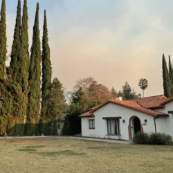 A white house with a red-tiled roof stands beside tall cypress trees on a lawn under a cloudy sky.