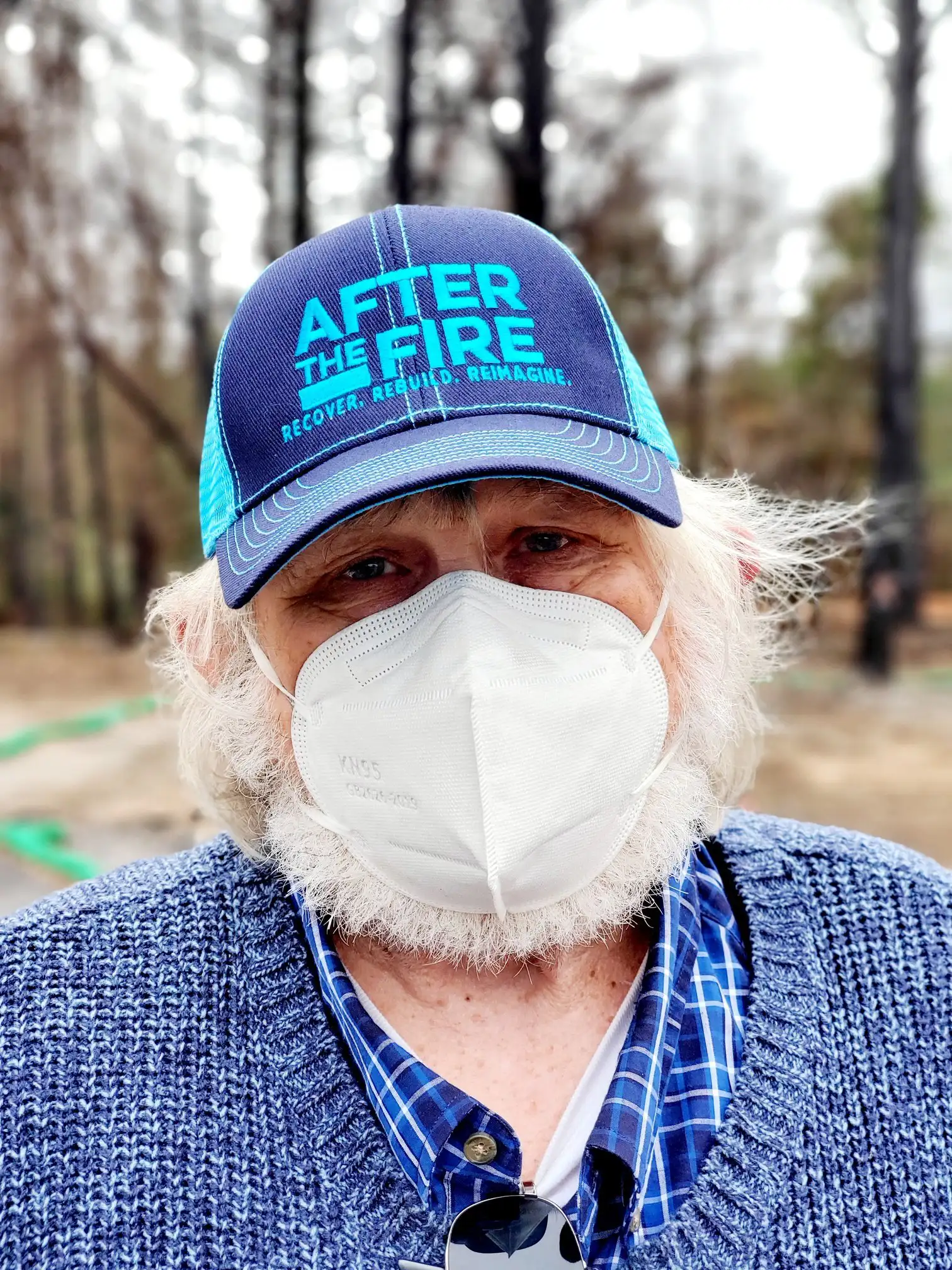 Older man in a mask and After the Fire hat stands outside, wearing a blue sweater and plaid shirt.