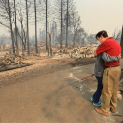 Two people hug on a street, surrounded by the burnt remains of trees and buildings after a fire.