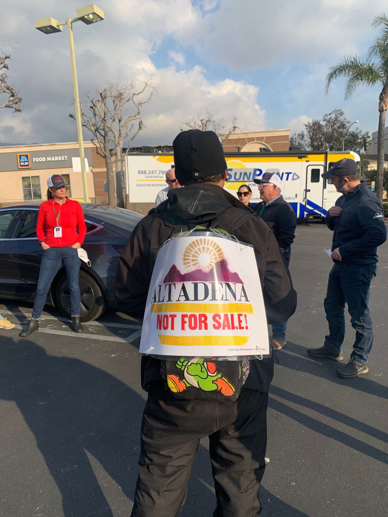 Person wearing a sign reading “Altadena Not For Sale!” stands in a parking lot with others talking nearby.