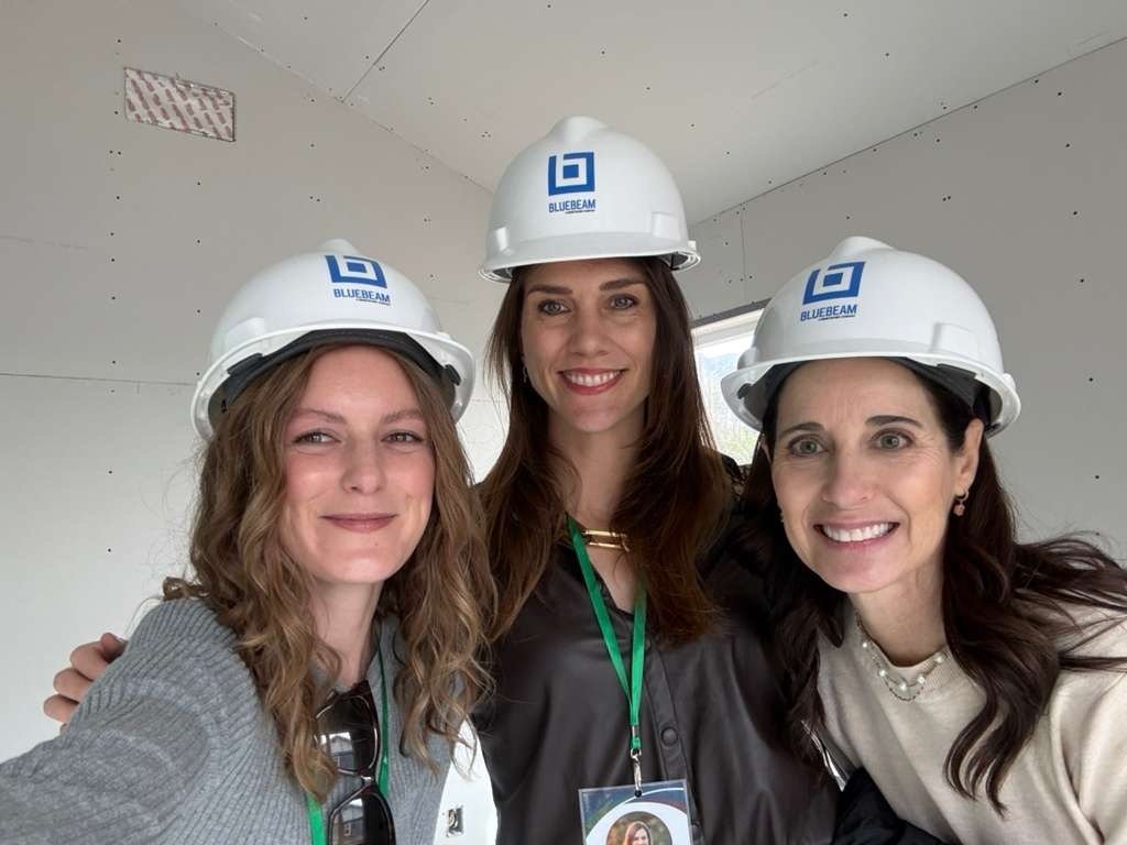 Three women wearing Bluebeam hard hats smile together indoors at a construction site.